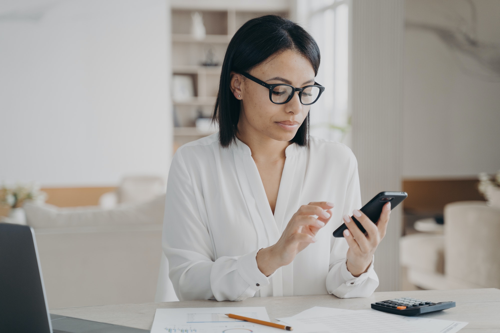 Femme d'affaires portant des lunettes et utilisant des applications de téléphonie mobile pour gérer son temps de travail, assise devant son ordinateur portable.