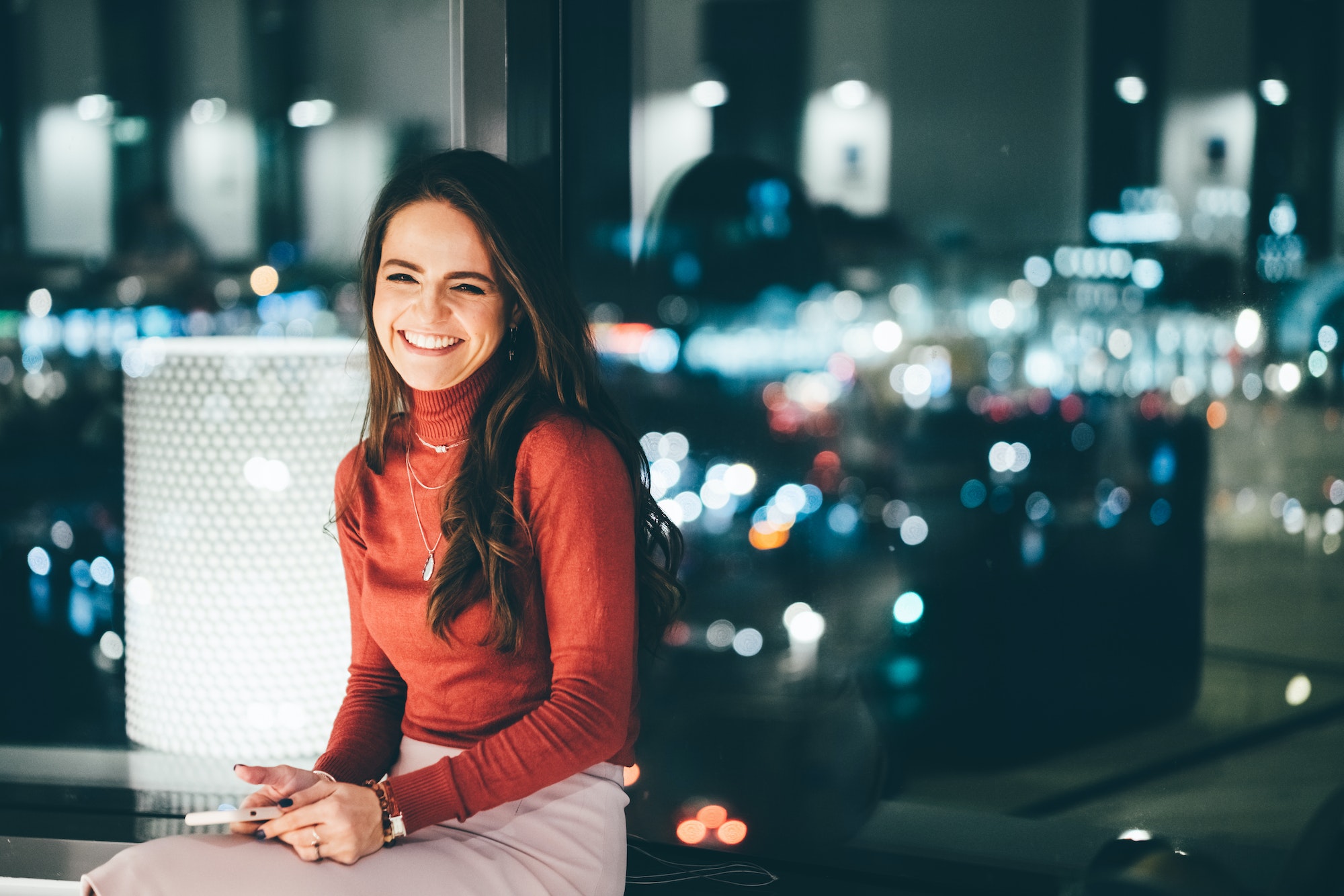 Portrait d'une jeune travailleuse utilisant son téléphone, regardant l'appareil photo et souriant sur son lieu de travail en soirée.